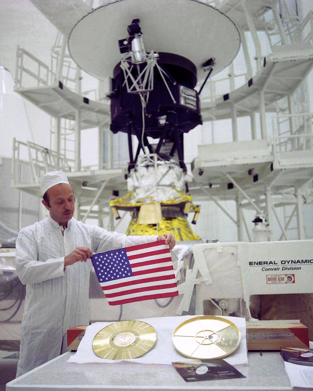 A medida que las dos sondas espaciales Voyager de la NASA viajan al espacio profundo, transportan una pequeña bandera estadounidense y un Disco de Oro repleto de imágenes y sonidos, recuerdos de nuestro planeta natal. Esta imagen muestra a John Casani, Gerente del proyecto Voyager en 1977, sosteniendo la pequeña bandera que fue doblada y cosida en las mantas térmicas de la nave Voyager antes de su lanzamiento. Debajo de Casani se puede ver el Disco Dorado (izquierda) y su cubierta (derecha). Al fondo se encuentra la sonda Voyager 2 antes de ser llevada a la plataforma de lanzamiento. La foto fue tomada en la base de Cabo Cañaveral, Florida (EUA), el 4 de Agosto de 1977. Crédito de la imagen: NASA / JPL-Caltech